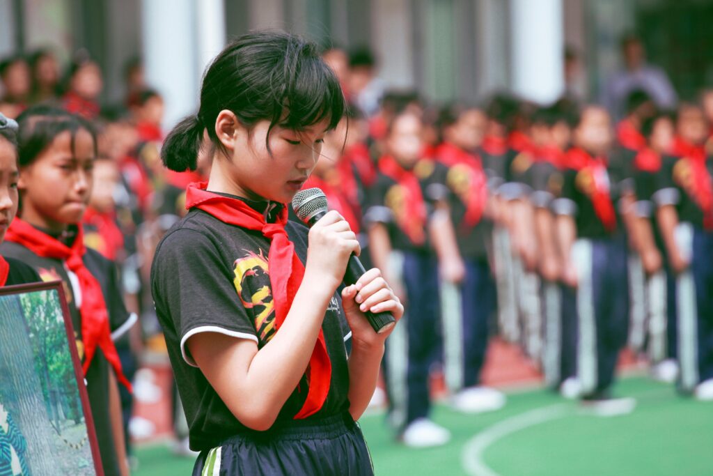 pupils-school-children-red-scarf-159671-159671 A young girl in uniform with a red scarf speaks into a microphone during an outdoor school event.